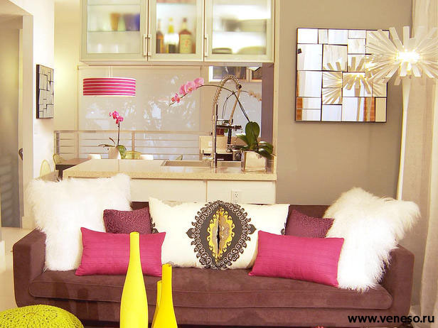 Living room with brown and pink decor, mirrored wall decor, fuzzy white pillows, soft couch, clear kitchen cabinets, and breakfast bar area.