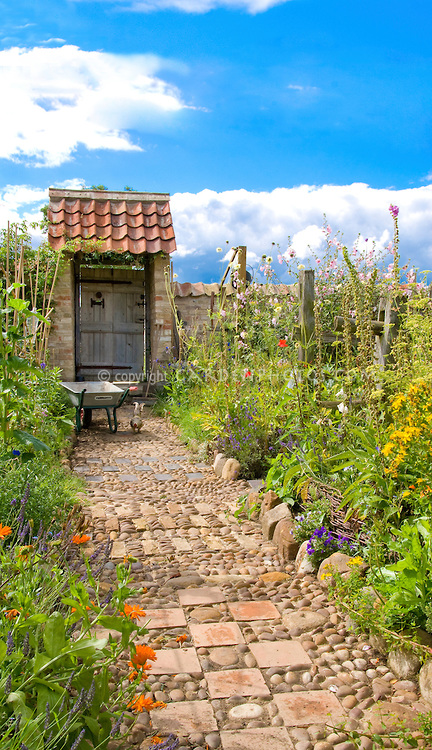 Garden path of stones, pebbles & tiles leading to garden gate door, with wheelbarrow, chicken, vegetable and flower garden borders, blue sky and clouds, wide view